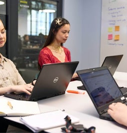 Two women on laptops during a team meeting with sticky notes in the background