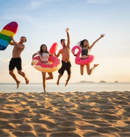 Four people in swimwear jump in the air on a sandy beach holding pool inflatables