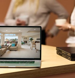 A modern laptop on a wooden table showcasing a virtual house tour with blurred individuals chatting over coffee in the background.