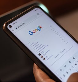 Hands holding a mobile device showing the Google homepage with a search query