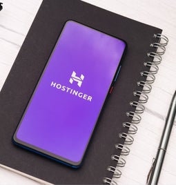 Cellphone shows the Hostinger logo, and sits upon a closed otebook next to glasses and a pen