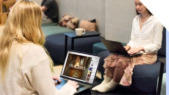 Two women using laptops in a modern office lounge area - one is watching an online service on YouTube.