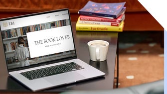 A comfortable reading setup with a laptop open on a blog about books, a cup of coffee, and a stack of books on a wooden table.