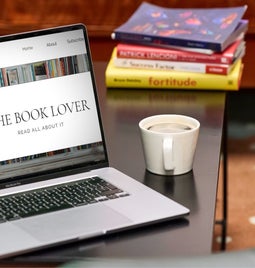 A comfortable reading setup with a laptop open on a blog about books, a cup of coffee, and a stack of books on a wooden table.