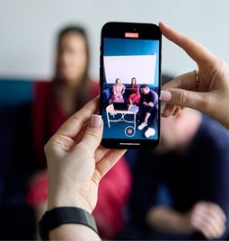 A close-up of a smartphone taking a video of friends for social media, with a blurred background.