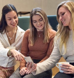 Three women smiling and looking at a smartphone together, with one woman pointing at something on the screen.