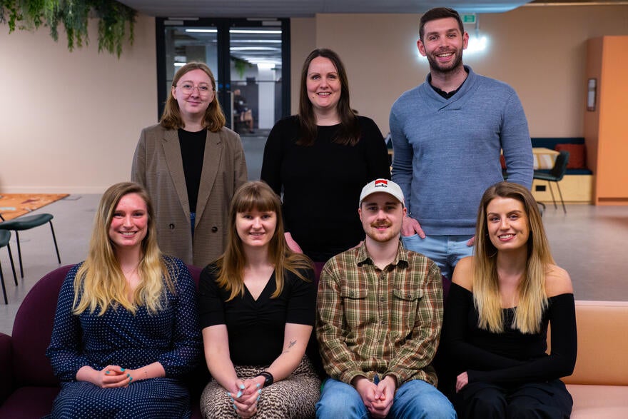 Seven members of the WBE team smile at the camera in an office